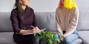 A person holding a clipboard smiles while sitting on a couch with a smiling teen. This symbolizes the support LGBTQIA+ therapy can offer. Contact a LGBTQ+ affirming therapist in North Carolina or search "lgbtq friendly therapist near me" to learn more today.
